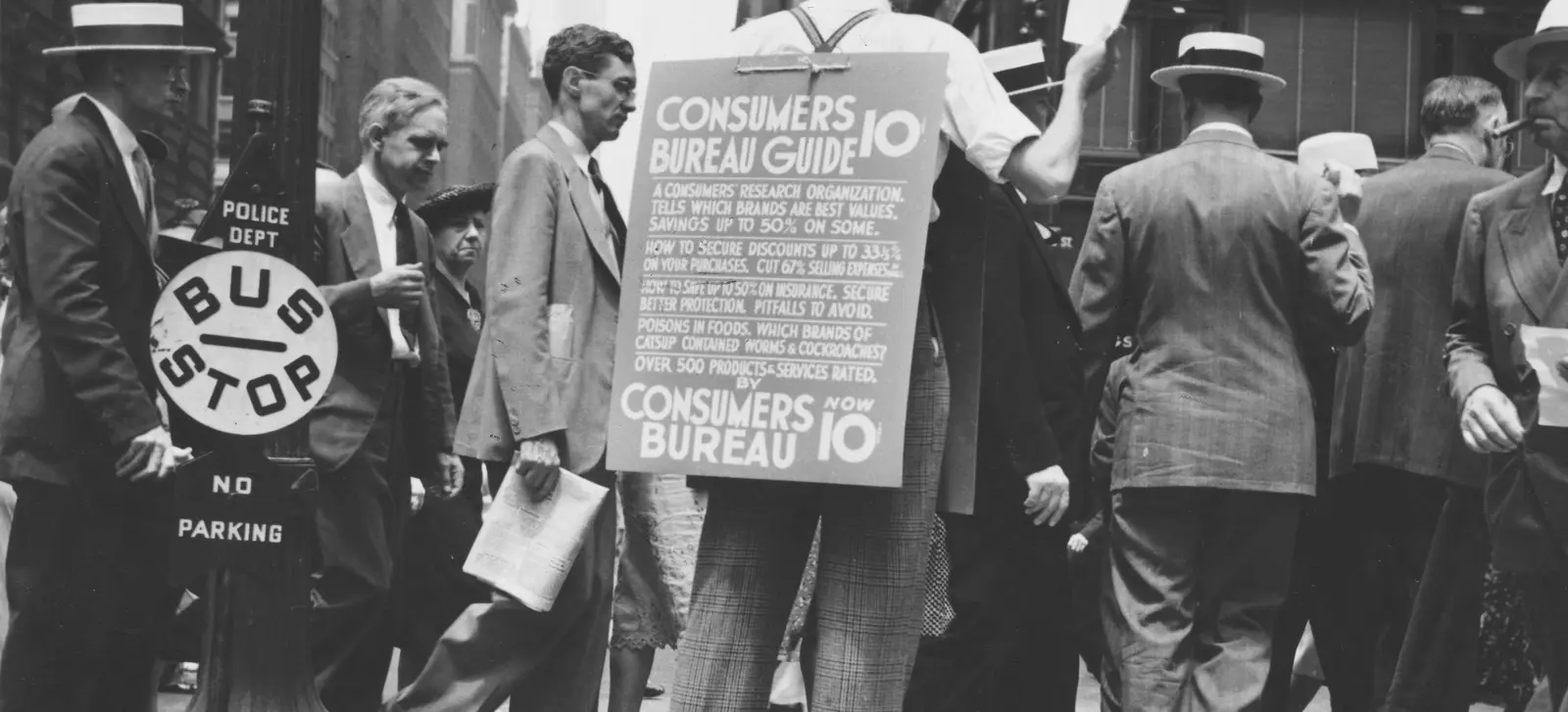 A man walking a crowded city street wearing a sandwich board advertising the Consumers Bureau Guide, a research organization rating over 500 products and services